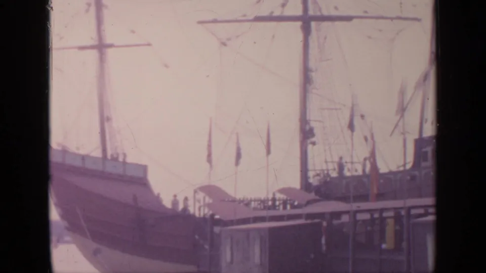 1964 Flushing Meadows home-movie frame showing a tall-masted display ship beside a small dock with flags and rigging.