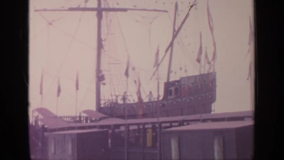 1964 Flushing Meadows home-movie frame showing the ship's deck area, tall mast, rigging, flags, and nearby dock structures.
