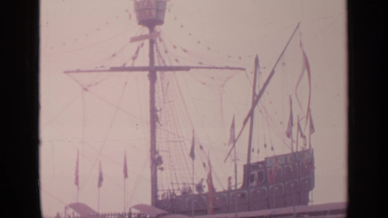 Tall ship model mast and crow's nest with rigging and flags against a pale sky.