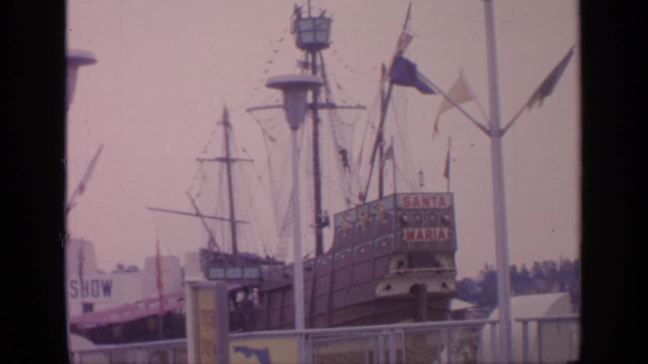 Santa Maria replica with masts, flags, fairground structures, and visitors behind a railing.