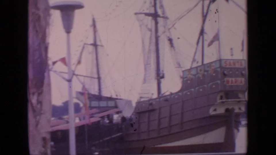 Handheld 1964 Flushing Meadows shot of the Santa Maria replica ship, showing the stern, rigging, flags, and nearby exhibit structures.