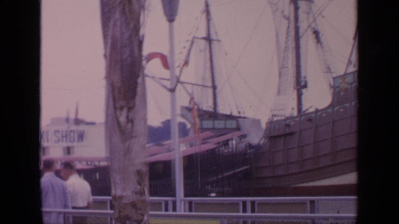 Visitors near railings with a large ship display and fairground structures in the background.