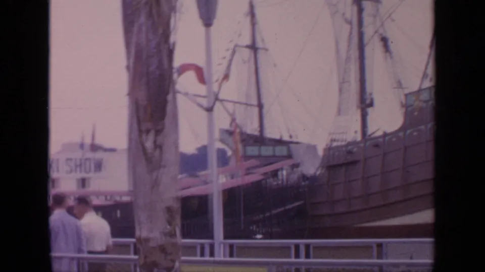 1964 Flushing Meadows home-movie frame showing visitors by railings, a tree trunk, and a large ship display on the fairgrounds.