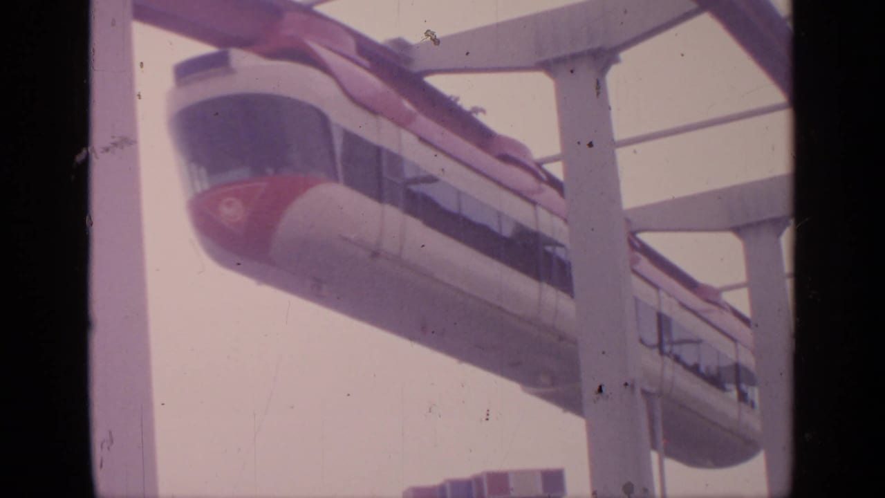 1964 Flushing Meadows home-movie frame showing a white-and-red monorail passing on an elevated guideway against a pale sky.