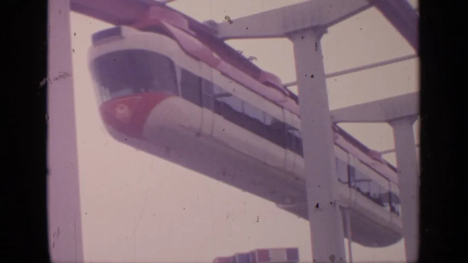 1964 Flushing Meadows home-movie frame showing a white-and-red monorail passing on an elevated guideway against a pale sky.