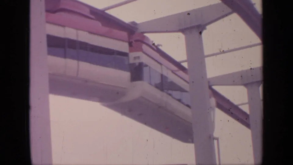 A low-angle 8mm frame shows the monorail car passing close to a white tower on the red guideway at Flushing Meadows.