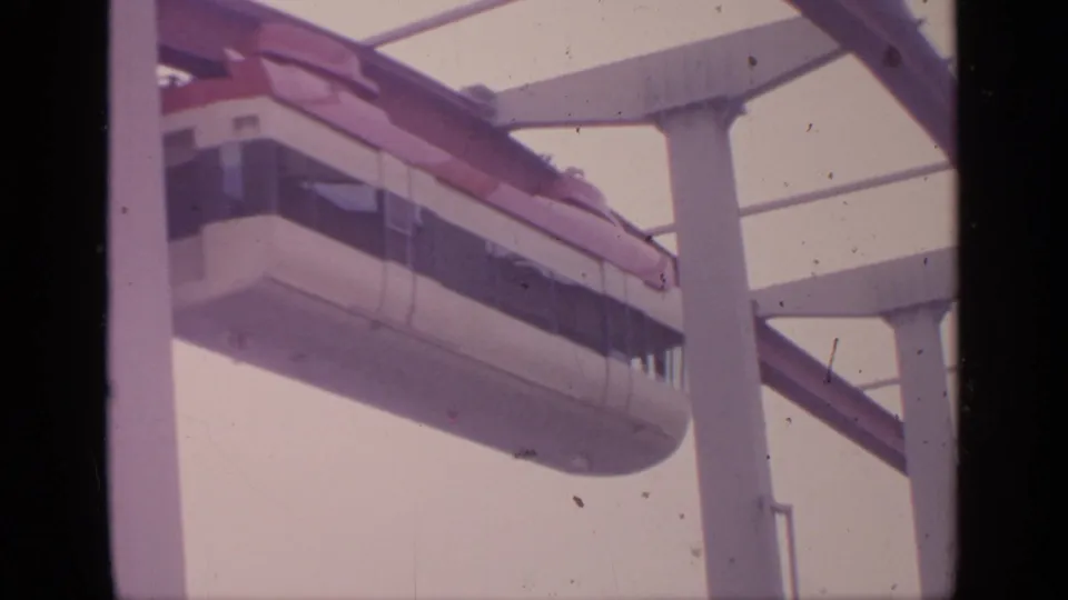 The monorail's trailing section moves through the frame beneath white supports in this 1964 Flushing Meadows home-movie still.