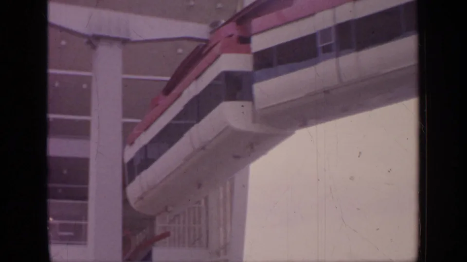A red-and-white monorail approaches a building at the 1964 Flushing Meadows setting, seen through soft 8mm home-movie footage.