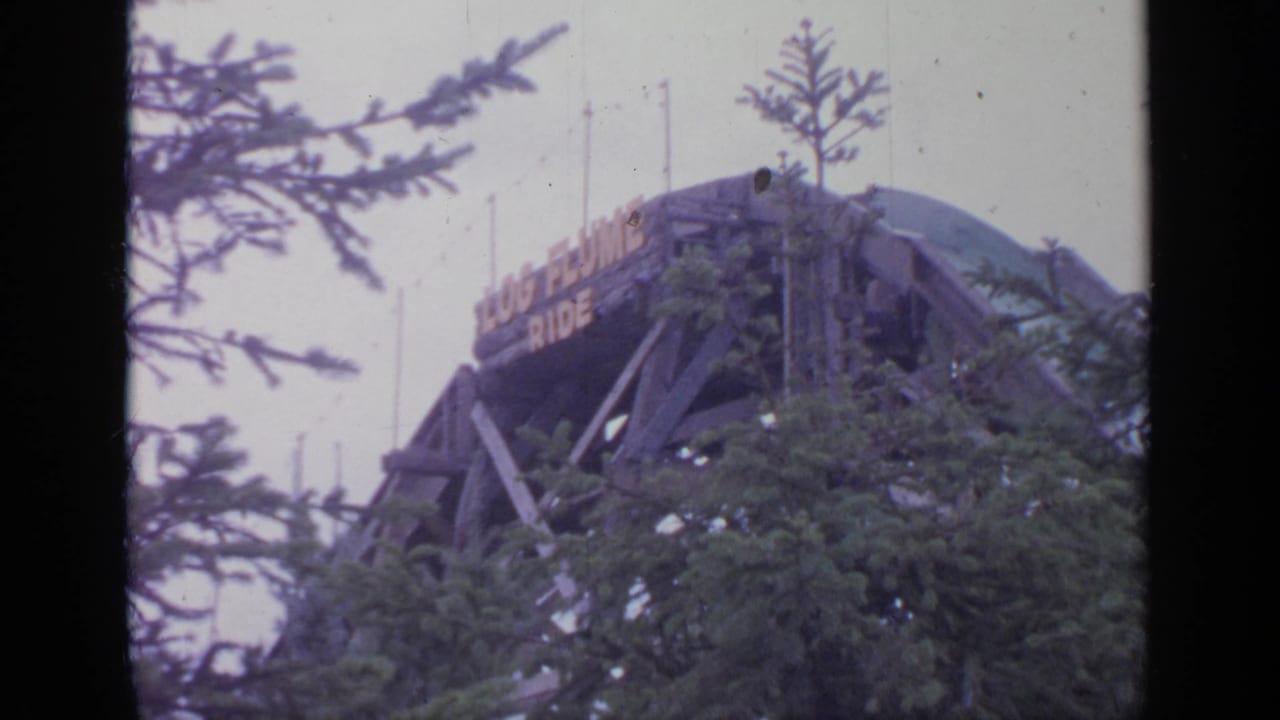 Flushing Meadows 1964 frame showing the upper wooden structure and sign of a log flume ride behind evergreen branches.