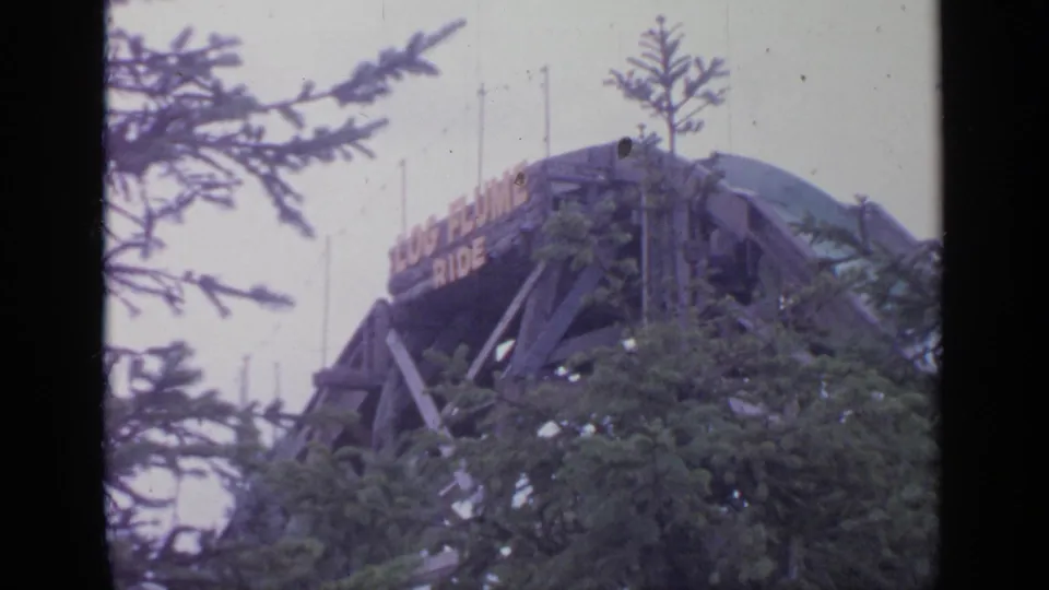 Flushing Meadows 1964 frame showing the upper wooden structure and sign of a log flume ride behind evergreen branches.