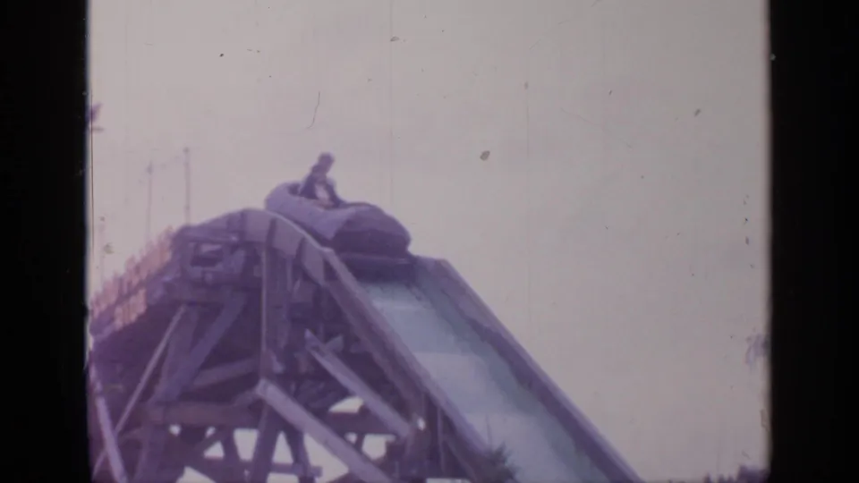 Visitor-shot 1964 frame of a log flume ride crest, with a small rider figure visible near the top of the chute.