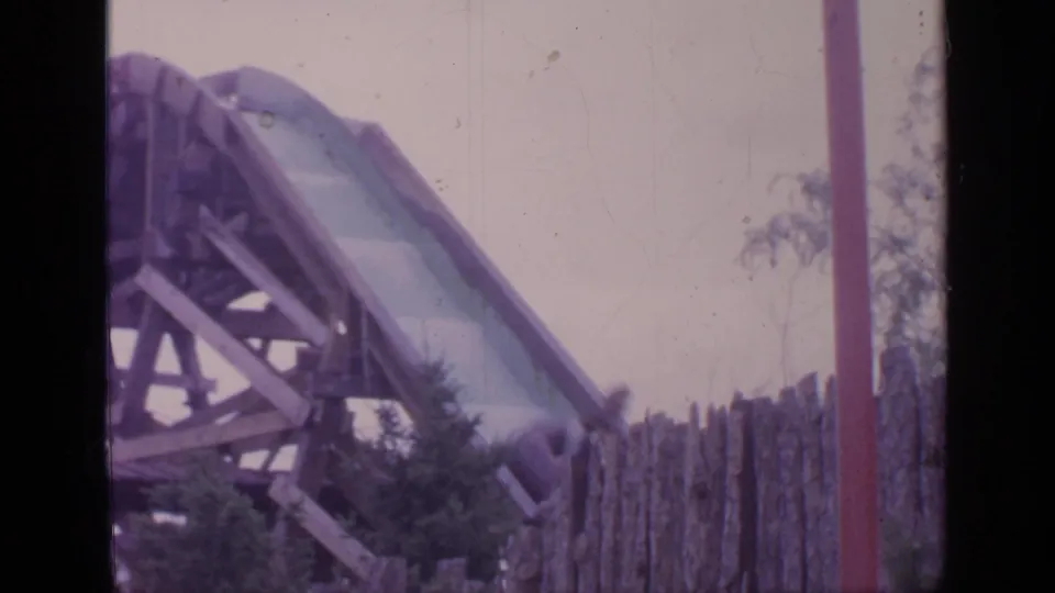 1964 Flushing Meadows frame showing the log flume chute descending past wooden supports, fencing, and nearby trees.