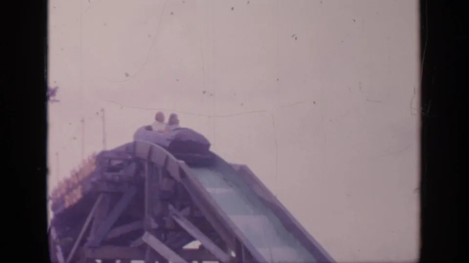 Two riders are visible in a coaster car cresting a steep drop at Flushing Meadows in 1964 8mm home-movie footage.