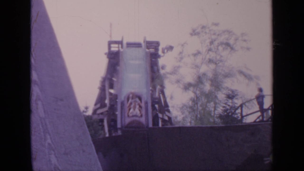 1964 Flushing Meadows frame with the log drop chute centered, wooden supports around it, trees behind, and a person standing near the platform railing.