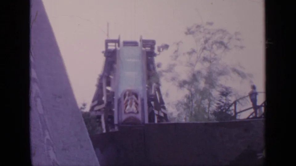1964 Flushing Meadows frame with the log drop chute centered, wooden supports around it, trees behind, and a person standing near the platform railing.