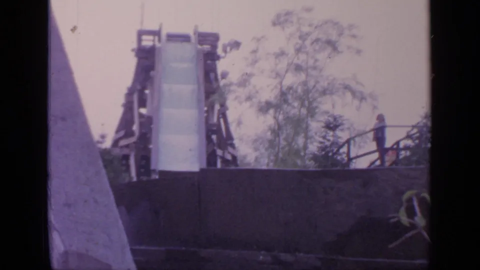 Visitor-shot 1964 Flushing Meadows frame showing the log ride chute, platform railings, trees, and a distant person moving along the walkway.