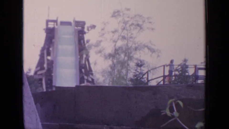 Final 1964 Flushing Meadows frame of the log drop ride, with the chute centered, trees behind it, and a distant person near the railing.