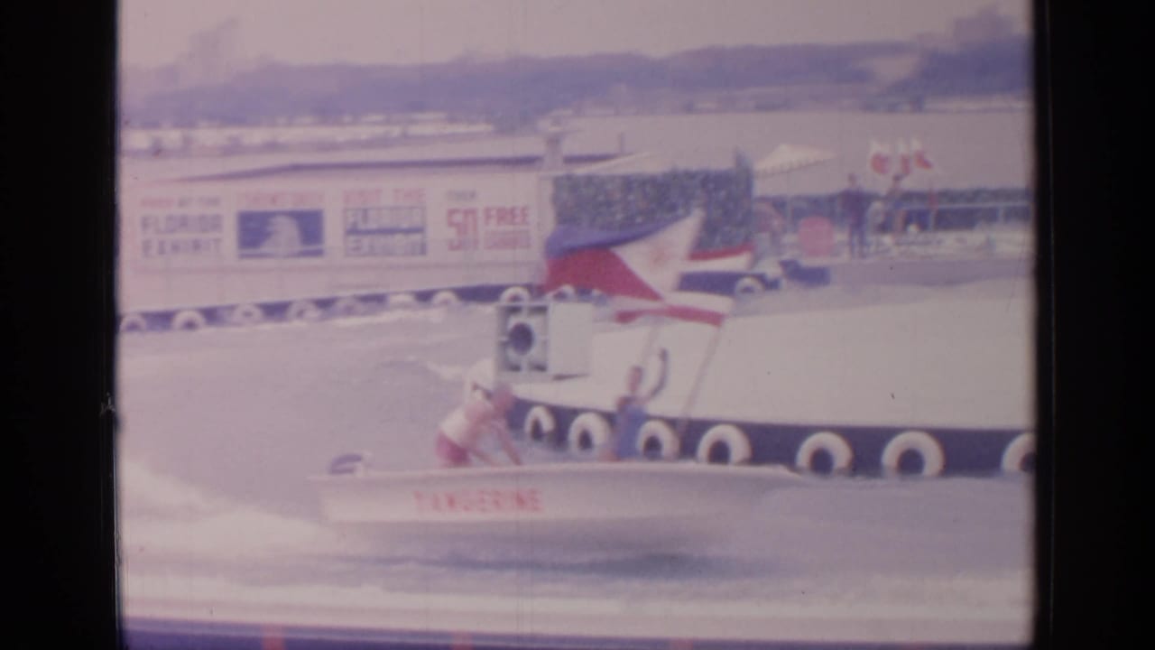 At Flushing Meadows in 1964, a speedboat marked Tangerine speeds past a platform and Florida Exhibit sign on a fairground water course.