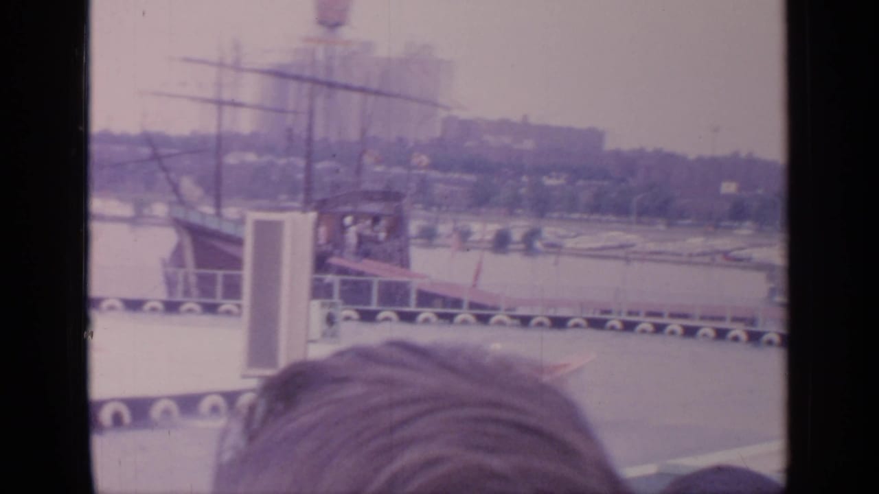 1964 Flushing Meadows World's Fair home-movie frame showing a water-stunt area, ramp, distant structures, and a spectator head in the foreground.