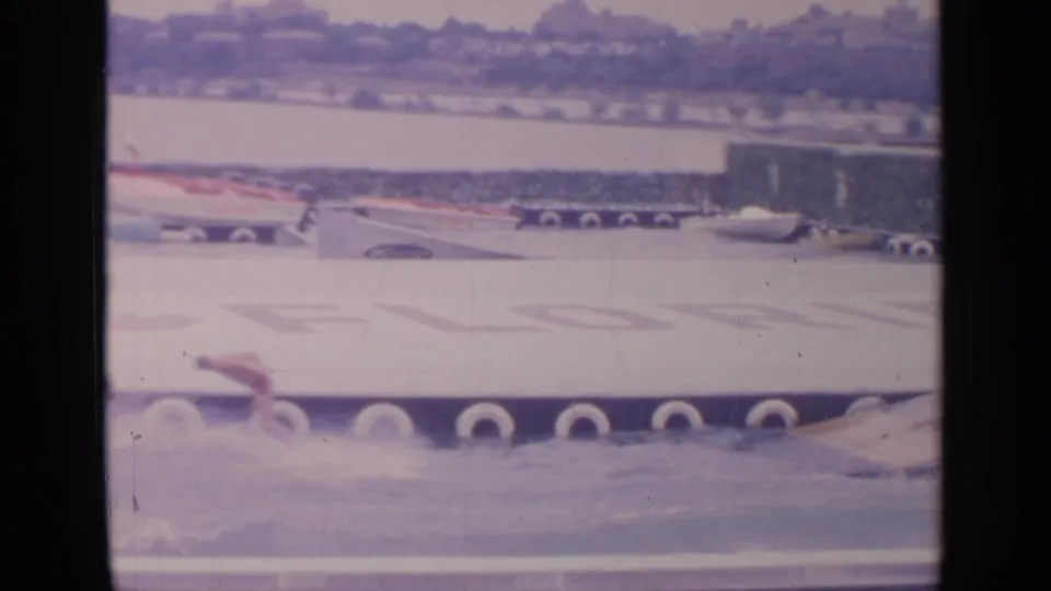 Home-movie frame from the 1964 Flushing Meadows World's Fair showing a water stunt with spray near the ramp and fair displays beyond.