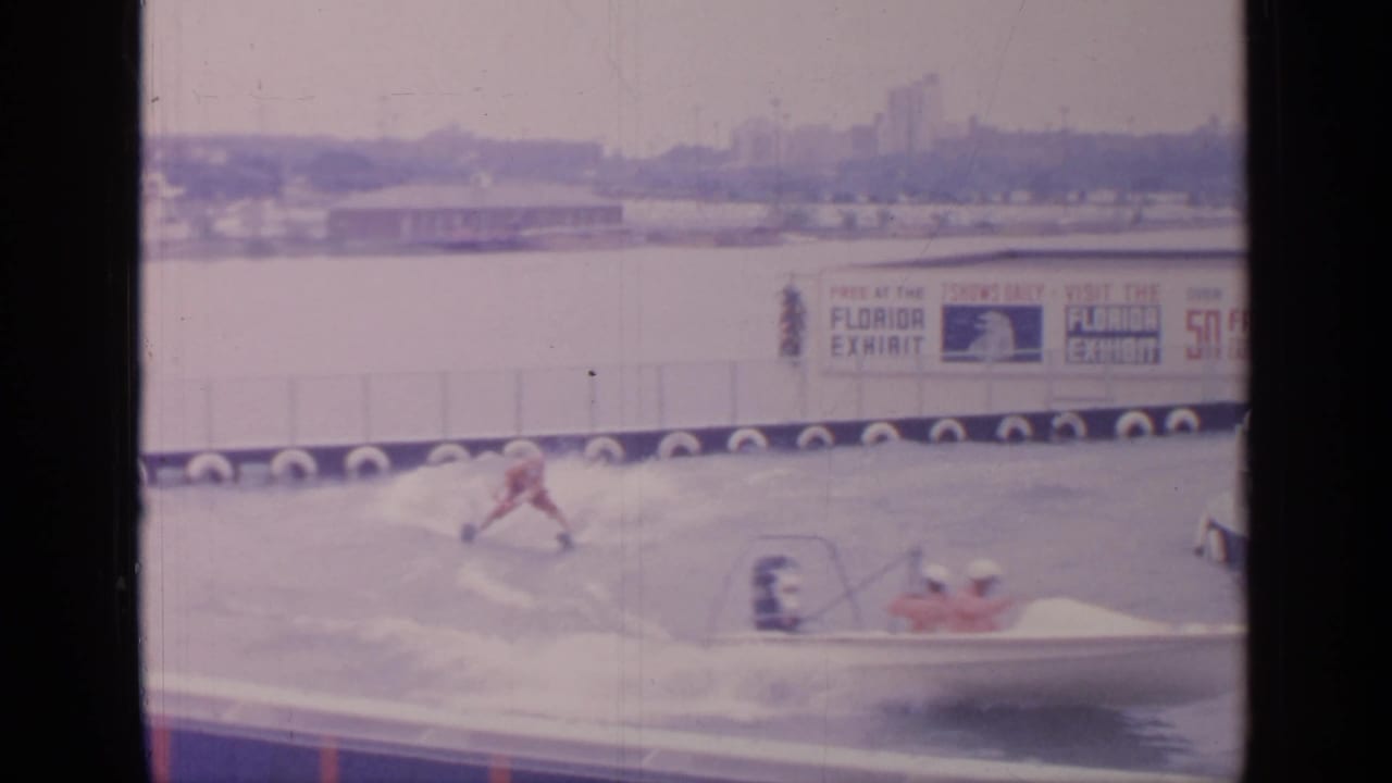 1964 Flushing Meadows home-movie frame showing a red-suited water skier, tow boat, spray, and Florida Exhibit signage in the background.