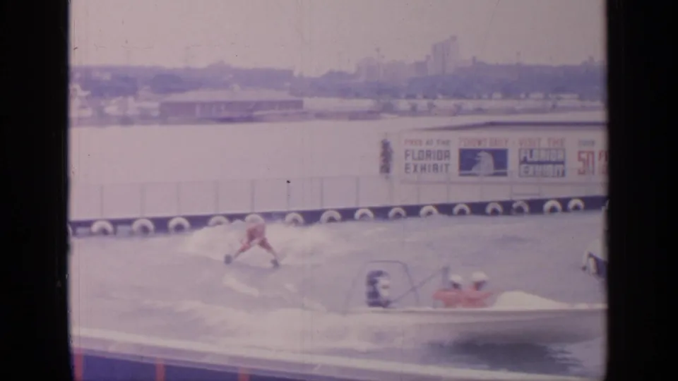 1964 Flushing Meadows home-movie frame showing a red-suited water skier, tow boat, spray, and Florida Exhibit signage in the background.