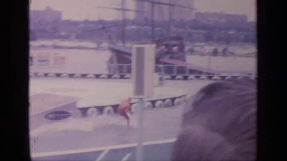 Home-movie view of the skier near the water's edge, partly framed by a foreground spectator and World's Fair structures beyond.