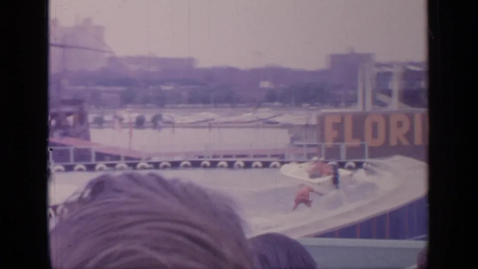Flushing Meadows frame with skier, tow boat, foreground spectators, lake barriers, and a large Florida Exhibit sign in the background.