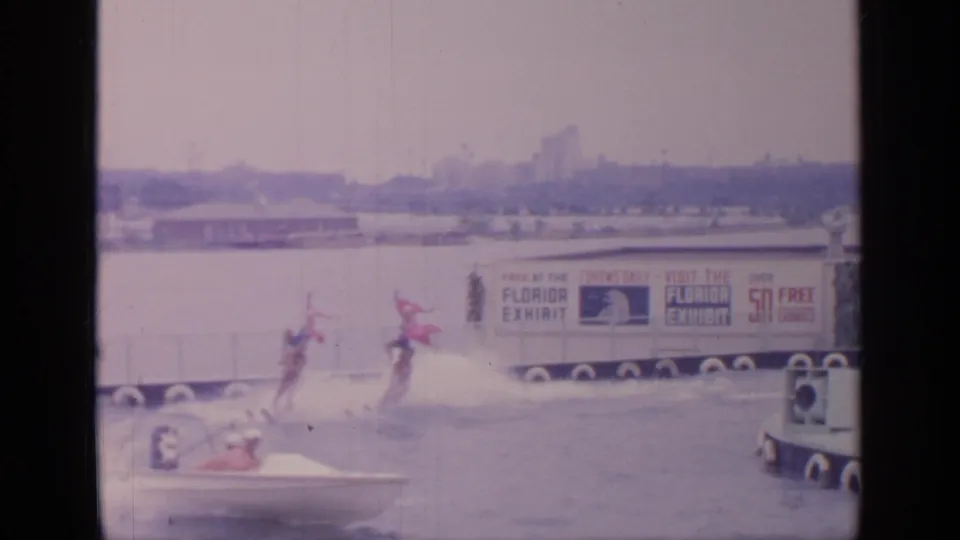Two skiers move across the water near a small boat and Florida exhibit banner in the 1964 Flushing Meadows fair footage.