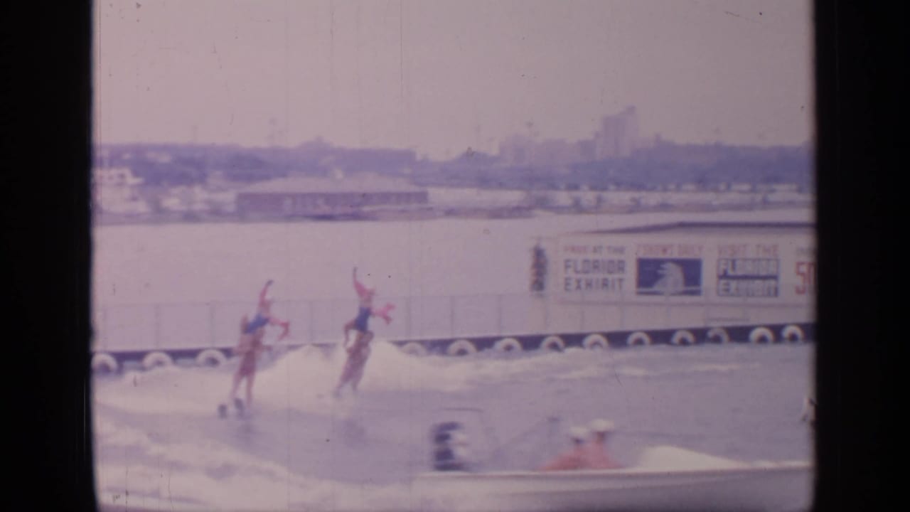 Two water skiers carrying red flags pass in front of a Florida Exhibit sign.
