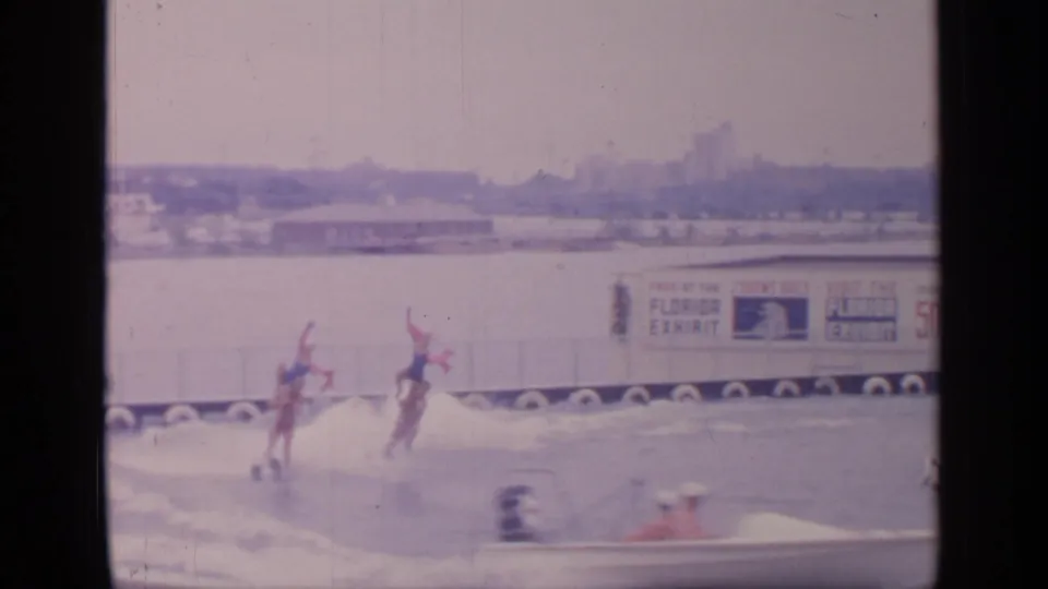 At the 1964 Flushing Meadows fair site, two water skiers with red flags move across the water near exhibit signage and barriers.