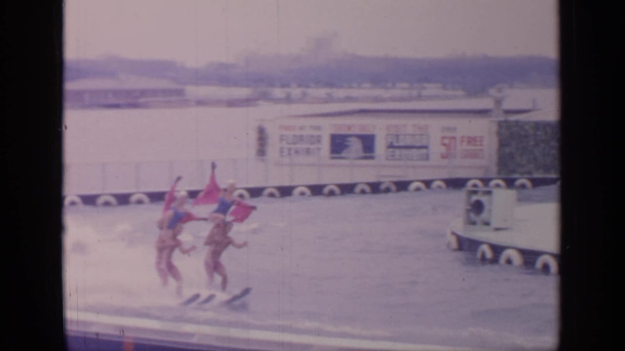Four water skiers move together across the water near exhibit buildings.