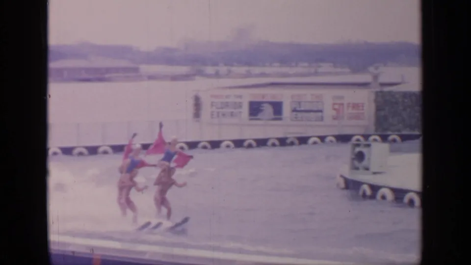 Four water skiers in coordinated formation pass the World's Fair waterfront area, with signage and a small tow craft nearby.