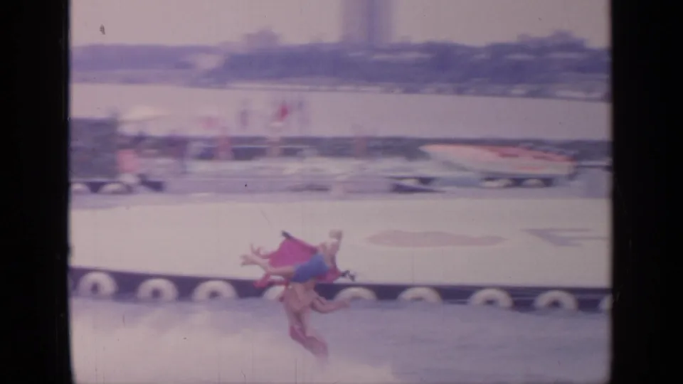 Two performers skim low over the water in a trick position, with a blurred boat and fair structures in the background.