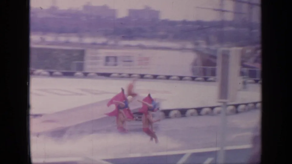 Two water skiers continue along the fairground waterway, partly blurred by motion, with exhibit structures and barriers behind them.