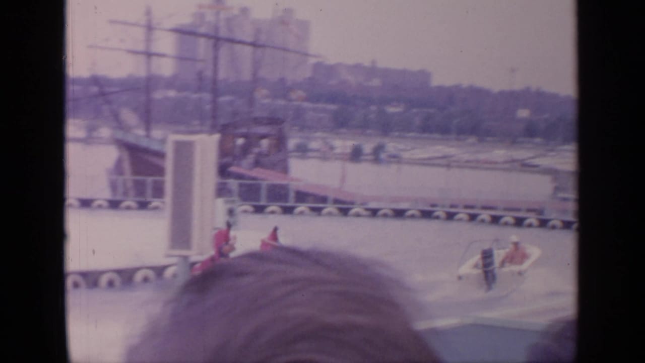 Waterskiers pass a fairground water stage as spectators' heads partially block the foreground.