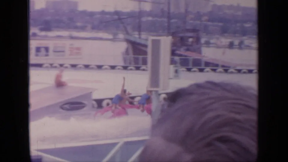 Home-movie frame of the water-ski act passing a white set column at Flushing Meadows, with spectators close to the camera.