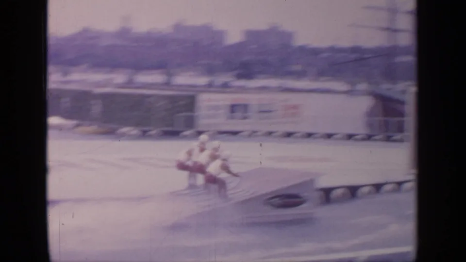 Two water skiers in 1964 Flushing Meadows footage pass near a floating ramp, with fairground signage and structures in the background.