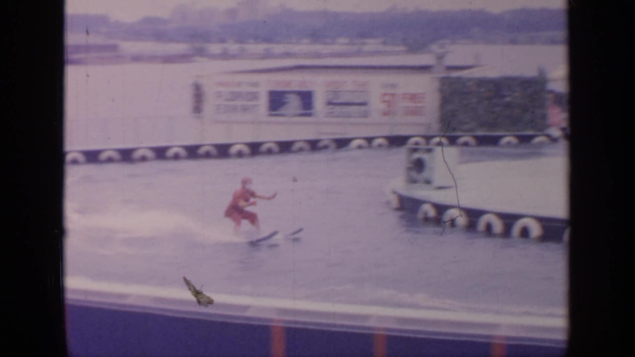 Waterskier sprays across the water while a floating platform and exhibit sign sit behind.