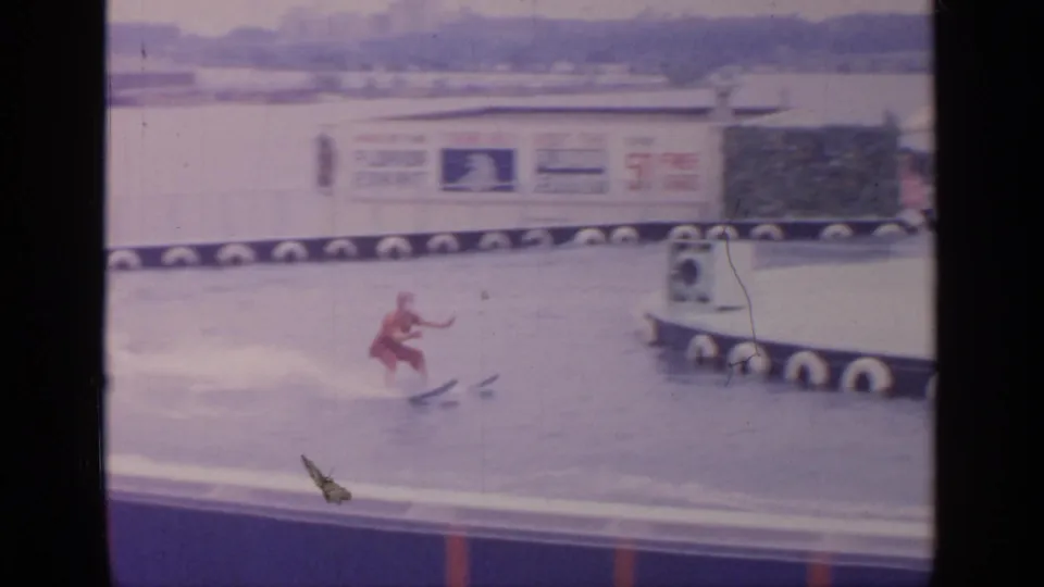 A red-clad waterskier glides past a floating platform at the 1964 Flushing Meadows fair site, with exhibit signage in the background.
