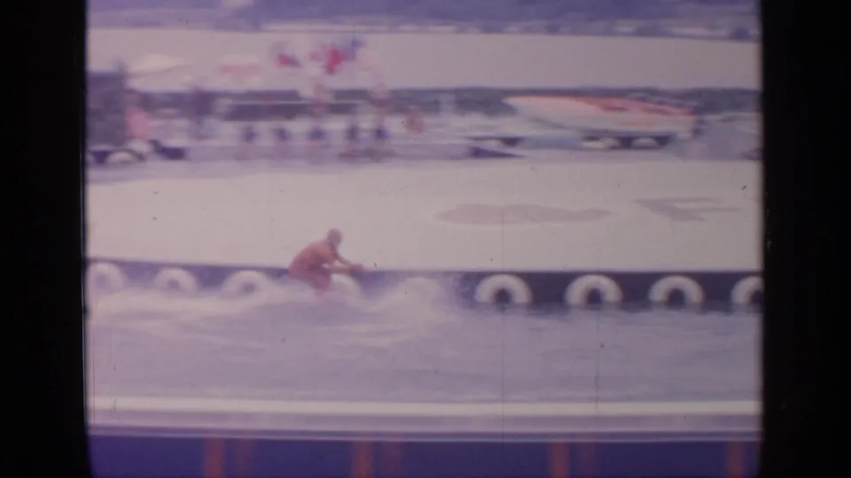 1964 Flushing Meadows footage shows the waterskier low on the water near a pier edge, with fairground structures beyond.