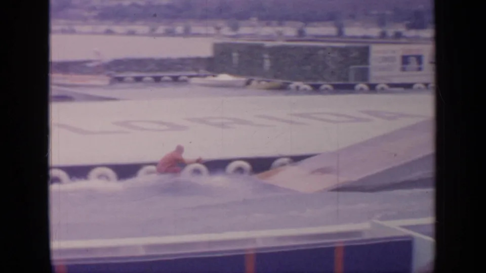The waterskier nears a ramp structure in the Flushing Meadows World's Fair water show area, with signage and pier barriers behind.