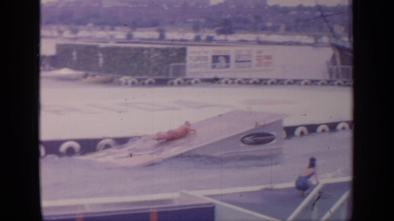 Water skier in red approaches a ramp with fairground signage across the water.