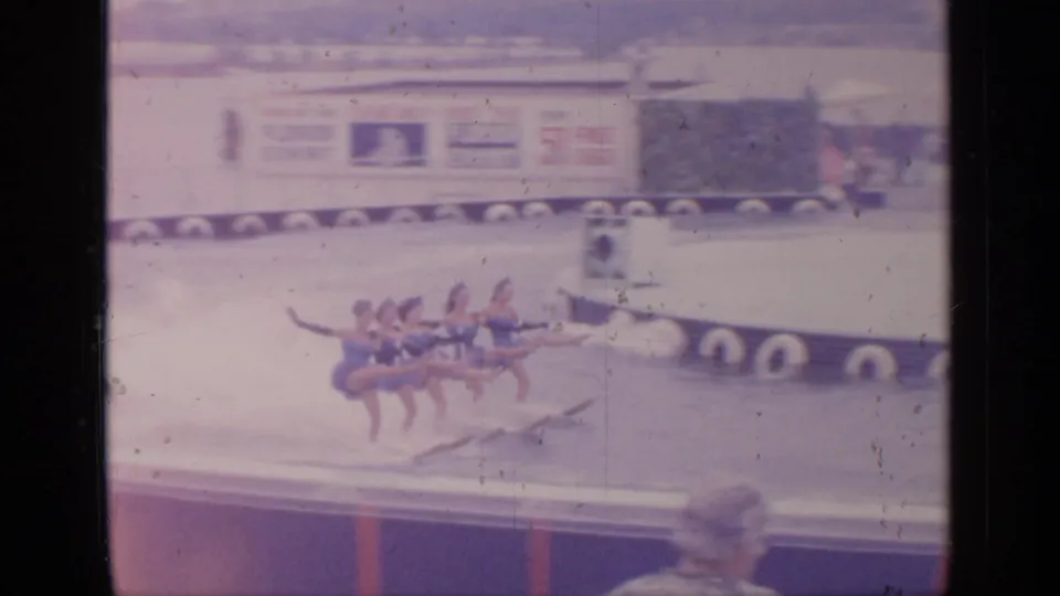 Female water skiers hold a formation on the water, with fair signage and spectators visible in the soft 8mm frame.