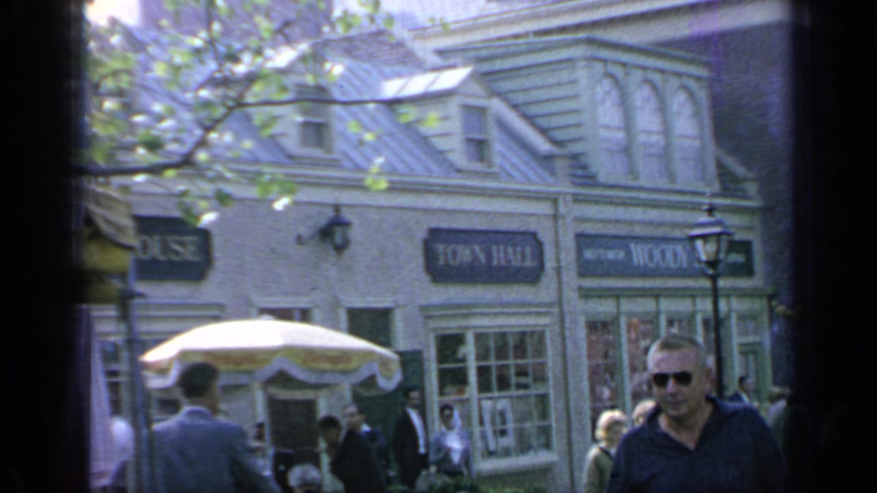 Visitors at the 1964 Flushing Meadows fair pass storefront facades with Town Hall and Woody's signs, umbrellas, and a lamppost.