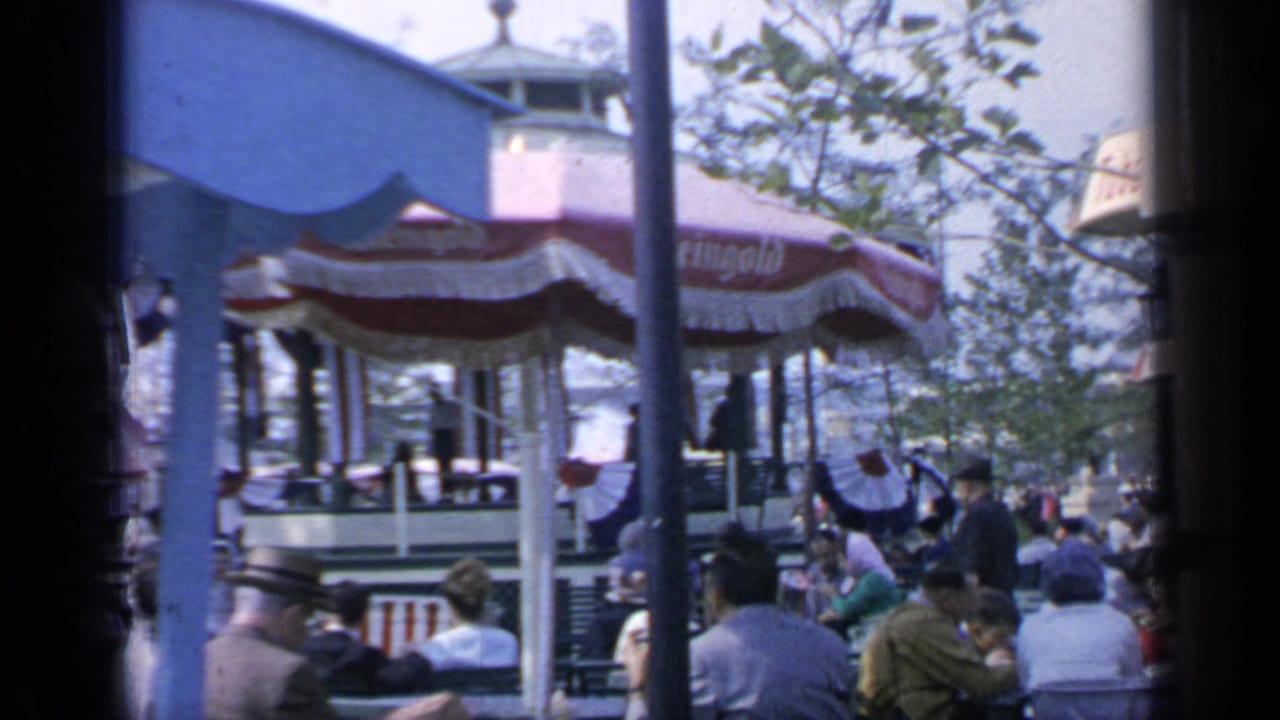Crowded 1964 Flushing Meadows outdoor seating area with striped concession umbrellas, trees, and visitors gathered beneath a blue canopy edge.
