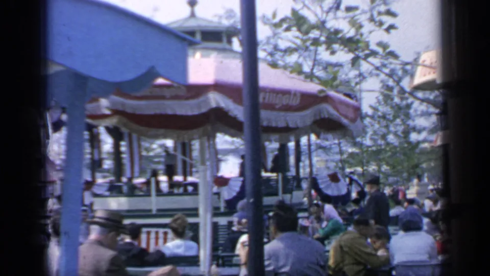 Crowded 1964 Flushing Meadows outdoor seating area with striped concession umbrellas, trees, and visitors gathered beneath a blue canopy edge.