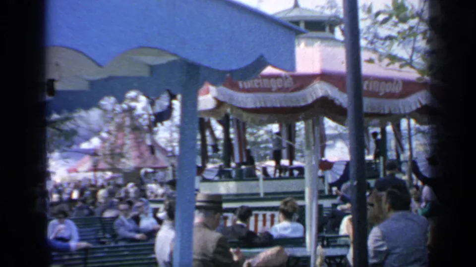 1964 Flushing Meadows crowd seated around benches and concession umbrellas, with fairground structures and trees visible beyond the blue canopy.