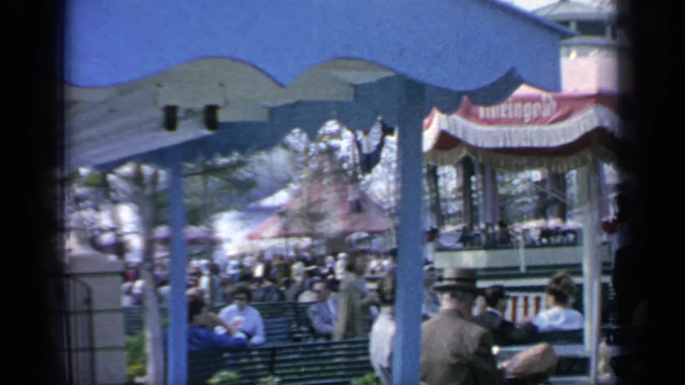 1964 Flushing Meadows visitors seated on benches near striped food umbrellas, with trees and fairground structures in the background.