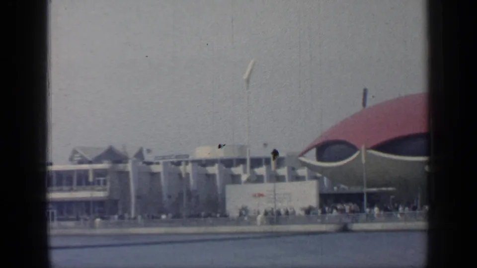 A panning 1964 fairground view shows modern structures, a partial red-domed pavilion, and distant visitors along the water at Flushing Meadows.